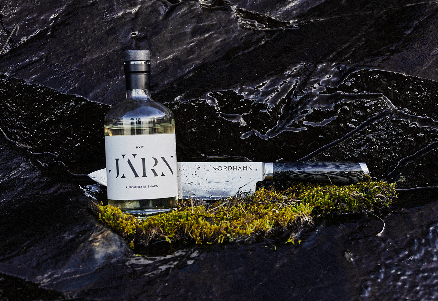 Kockkniv från Nordhamn mot svart granit och en bädd av mossa. Tillsammans med kniven från en flaska alkoholfri snaps från Järn Brands.