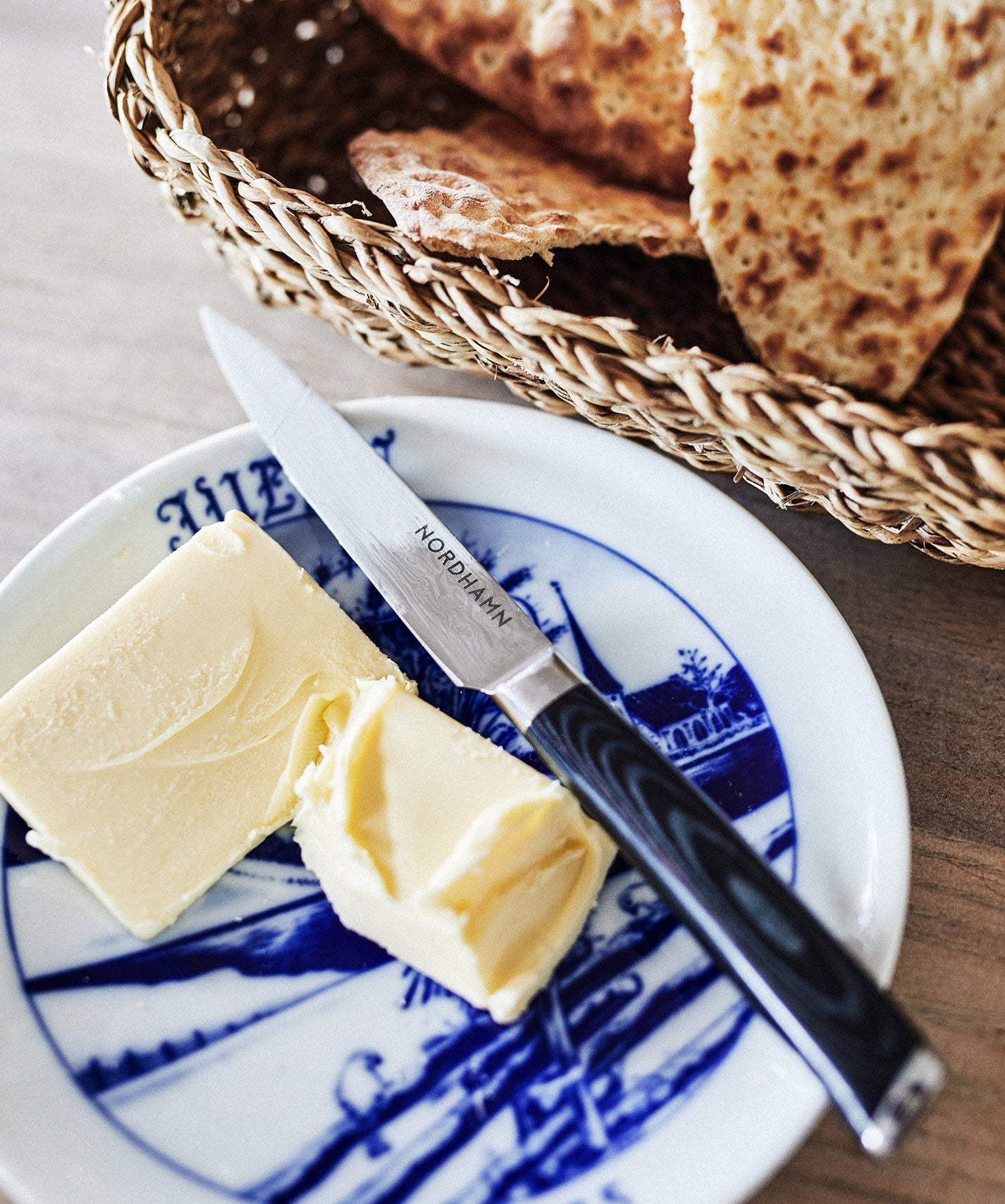 Allroundkniv från Nordhamn ligger på en gammal julassiett tillsammans med två klickar smör. Bredvid står en flätad korg med tunnt bakad dyrökaka.